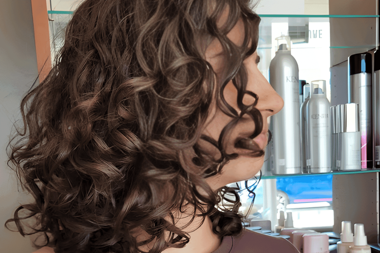 Curly-haired person with salon products on a shelf in the background.